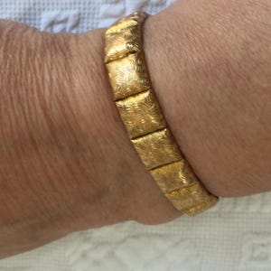 May include: A gold-toned bracelet with a repeating rectangular pattern. Each rectangle features an etched, swirling design. The bracelet is worn on a wrist, and the clasp is not visible.