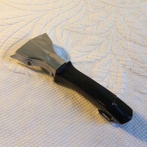 May include: A metal pan scraper with a black handle. The scraper has a wide, angled blade and a black, ergonomic handle with a small metal loop. The blade is silver and appears to be made of stainless steel.