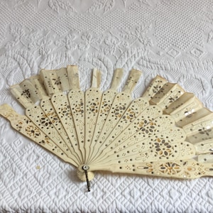Puede incluir: Un abanico de mano antiguo de color marfil, abierto y expuesto. El abanico presenta intrincados patrones florales y de puntos en marrón y dorado. El abanico está hecho de un material fino y delicado y tiene un pequeño cierre de metal. El abanico está sobre una superficie blanca y texturizada.