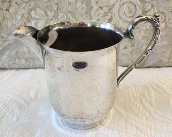 May include: A silver-colored pitcher with a curved handle and spout. The pitcher has a rounded body and a decorative pattern on the surface. The handle is ornate, and the spout is designed for easy pouring. The pitcher is likely made of metal and is suitable for serving beverages.