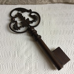 May include: A decorative, oversized key with a dark brown finish. The key's head features an ornate design with leaf and scroll details. The shaft and bit are rectangular. The key is approximately 12 inches long.