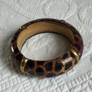 May include: A round bangle bracelet with a brown and black animal print design. The bracelet has gold-colored metal accents and a light brown interior. The bracelet is made of wood and has a glossy finish.