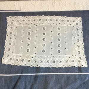 May include: A white rectangular doily with an intricate eyelet design. The doily features a scalloped edge and a pattern of small floral and circular cutouts. The doily is placed on a blue and white striped surface.