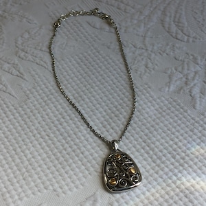 May include: A silver-toned chain necklace featuring a teardrop-shaped pendant with an openwork scroll design. The pendant is accented with small, smooth, gold-toned circular elements integrated into the intricate metalwork of the frame.