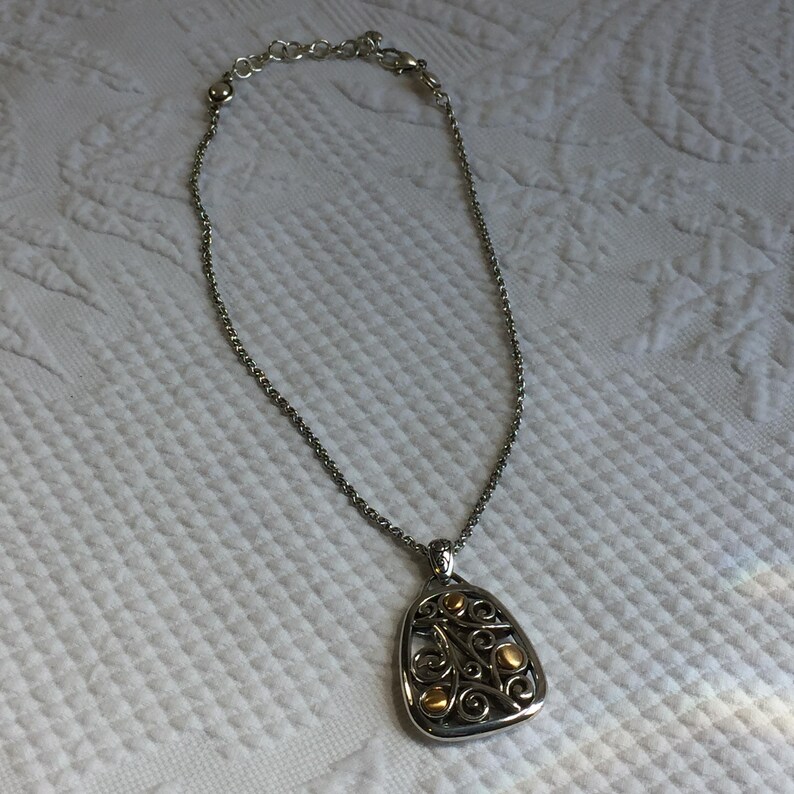 May include: A silver-toned chain necklace featuring a teardrop-shaped pendant with an openwork scroll design. The pendant is accented with small, smooth, gold-toned circular elements integrated into the intricate metalwork of the frame.