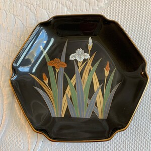 Può includere: Un piatto ottagonale in ceramica nera con un bordo dorato e un motivo floreale con iris bianchi, arancioni e verdi su sfondo nero.