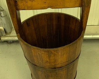 Puede incluir: Un cubo de madera vintage con asa, hecho de madera natural con un acabado marrón oscuro. El cubo está sentado en una caja blanca con una tapa rosa.