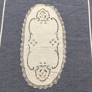 May include: Oval white linen doily with detailed cutwork and a fine lace trim. The design incorporates floral and geometric motifs, offering a vintage and elegant appearance. The doily is displayed on a blue and white striped background.