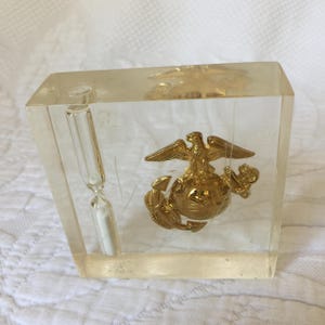 May include: A clear acrylic block with a gold-colored United States Marine Corps emblem inside. The emblem features an eagle perched on a globe with an anchor. A small hourglass is also visible inside the block.