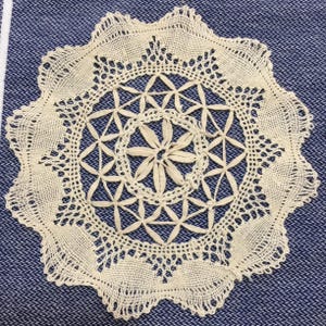 May include: Cream-colored, handmade doily with a floral and geometric design. The doily features a central flower motif surrounded by star-shaped patterns and a scalloped edge. The intricate lace is set against a blue fabric background.
