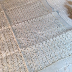 May include: Close-up of a white lace fabric with a floral pattern. The delicate lace is laid out, showcasing the intricate design and texture. The fabric appears to be a tablecloth or decorative textile, with a soft, off-white color.