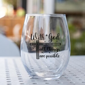 May include: A clear stemless wine glass with the text "With God all things are possible" and a cross design in black. The text includes the Bible verse Matthew 19:26. The glass is on a white surface.