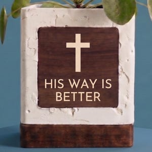 May include: A square, white and brown planter with the text "HIS WAY IS BETTER" and a cross. The planter has a wooden panel in the center with the text and cross. Green plant leaves are visible at the top.