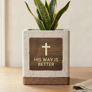 May include: A square, white planter with a wooden base and a wooden plaque featuring a cross and the text "HIS WAY IS BETTER." The planter holds a snake plant with green and yellow leaves. The planter is on a wooden surface.