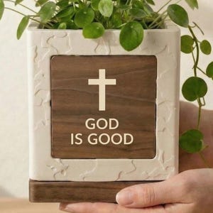 May include: A square, off-white planter with a wooden panel featuring a white cross and the text "GOD IS GOOD." The planter holds a green plant with round leaves. The planter is being held by a person.