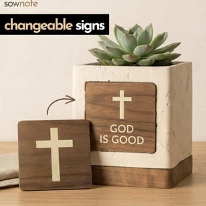 May include: A square, cream-colored planter with a wooden base and a succulent plant. A wooden sign with a white cross and the words "GOD IS GOOD" is inserted into the planter. A separate wooden sign with a white cross is next to the planter. The text "changeable signs" is above the planter.