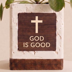 May include: A rectangular planter with a white textured frame and a dark wood-grain center. The center features a cross and the words "GOD IS GOOD" in a light beige color. The planter holds a green plant.