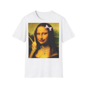 May include: White t-shirt with a pop art image of the Mona Lisa. The Mona Lisa is wearing a pink bow, a pearl necklace, and is making a peace sign. A humorous take on the classic painting, suitable for art lovers.