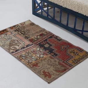 Può includere: Un tappeto rettangolare patchwork con una varietà di colori e motivi. Il tappeto presenta un mix di tonalità rosse, beige e marroni, con disegni geometrici e floreali. Sullo sfondo c'è una panca blu con un cuscino bianco.