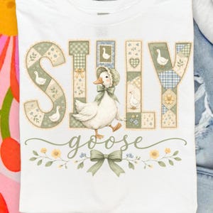 May include: White t-shirt featuring the words "SILLY GOOSE" in a patchwork design with a cartoon goose wearing a bonnet. The letters are in shades of green, blue, and beige, with floral and goose accents. A green bow and floral design are at the bottom.