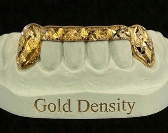 Puede incluir: Una parrilla dorada colocada sobre un molde dental blanco. La parrilla tiene un diseño texturizado y cubre cuatro dientes. La base del molde tiene las palabras "Gold Density" en letras doradas.