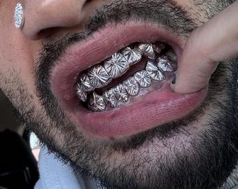 Puede incluir: Primer plano de la boca de una persona con grillz plateados. Los grillz presentan un patrón de estrella repetido. Se ve un collar de cadena plateada, junto con un pequeño pendiente de diamante.