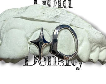 Puede incluir: Grillz dentales plateados con una estrella y una forma redondeada, exhibidos en un molde dental blanco. El texto "Gold Density" está superpuesto en la imagen en negro.