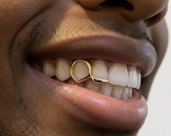 Puede incluir: Primer plano de una sonrisa, mostrando una rejilla dental dorada. La rejilla es personalizada, con una superficie pulida y reflectante. Los dientes son blancos, contrastando con el oro. El tono de piel es oscuro.