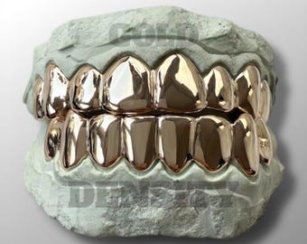 Puede incluir: Un juego de grillz dorados, exhibidos sobre un molde dental verde claro. Los grillz están muy pulidos, reflejan la luz y están diseñados para cubrir los dientes superiores e inferiores. Las palabras "GOLD DENSITY" son visibles en el molde.
