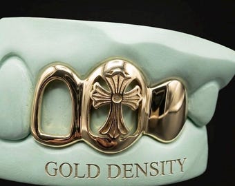 Puede incluir: Una parrilla dorada con un diseño de cruz, exhibida en un molde dental verde claro. El texto "GOLD DENSITY" está grabado en el molde. La parrilla tiene una superficie brillante y reflectante.