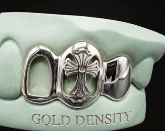 Puede incluir: Un grill dental de color plateado con un diseño de cruz recortada sobre un molde dental verde claro. El molde tiene grabado el texto GOLD DENSITY. El grill cubre tres posiciones dentales con un acabado metálico pulido y detalles intrincados.