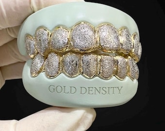 Puede incluir: Un juego de grillz dentales dorados con apariencia de polvo de diamantes, colocados sobre un molde verde claro. El texto "GOLD DENSITY" está grabado en el molde. Los grillz se muestran sobre un fondo oscuro.