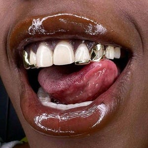 Può includere: Primo piano di una bocca con labbra lucide marrone scuro e denti bianchi. Grillz dorati con disegni a forma di stella sono visibili sui denti superiori. La lingua è estesa e l'estetica generale è glamour.