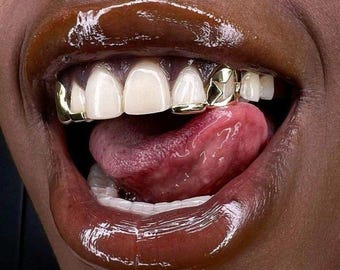 Puede incluir: Primer plano de una boca con labios brillantes de color marrón oscuro y dientes blancos. Se ven grillz dorados con diseños en forma de estrella en los dientes superiores. La lengua está extendida y la estética general es glamurosa.