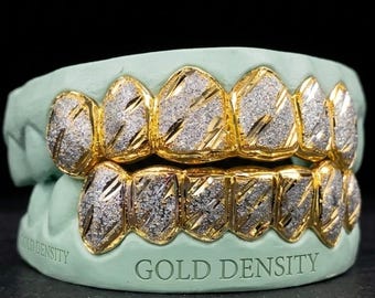 Puede incluir: Grillz de oro con apariencia de polvo de diamantes, colocados sobre un molde verde claro. Los grillz tienen un marco dorado con un patrón diagonal texturizado. El texto "GOLD DENSITY" es visible en el molde.