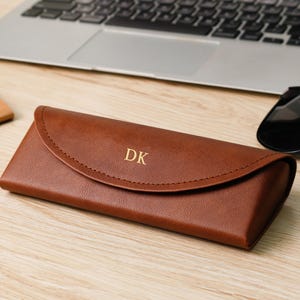 Pode incluir: Um estojo de óculos de couro castanho com as iniciais "DK" em letras douradas. O estojo está fechado e está sobre uma superfície de madeira ao lado de um par de óculos de sol e um laptop.