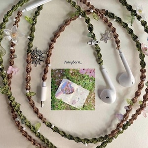 May include: Earphone cord with a macrame design in shades of brown and green, adorned with clear plastic flowers, leaves, and silver charms. The cord has white earbuds and a white charging port. The text "fairyborn_" is visible.