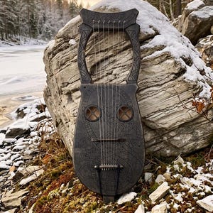 May include: A dark gray lyre with intricate carvings and strings, set against a snowy rock and a winter landscape. The instrument features a unique design with circular sound holes and decorative patterns, evoking a sense of history and craftsmanship.