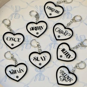 May include: Heart-shaped keychains with black borders and silver key rings. Each keychain features a different word in white, including "ONCE", "ARMY", "CARAT", "SLAY", "ATINY", "BLINK", "ENGENE", and "VILLAIN". A small asterisk is below each word.