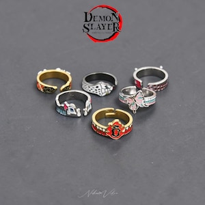 May include: A selection of seven adjustable rings with vibrant enamel designs. The rings showcase diverse patterns, including a butterfly, floral, and geometric shapes. The rings are silver and gold-toned, with the text "Demon Slayer" in the top right corner.
