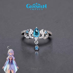 May include: A silver ring with a central light blue teardrop-shaped gemstone, surrounded by smaller blue stones and clear crystals. The ring has an intricate design with a dangling blue gem. The text "GENSHIN IMPACT" is displayed above the ring.