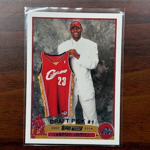 May include: A Topps rookie card featuring LeBron James holding a Cleveland Cavaliers jersey. The card is labeled "Draft Pick #1" and includes the years 2003 and 2004. The jersey is red with gold accents and the number 23.
