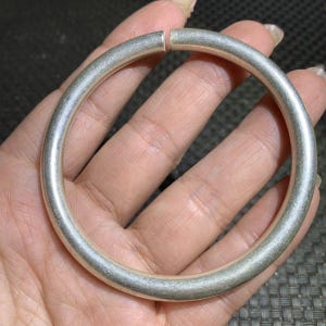 Może przedstawiać: Srebrna, okrągła bransoletka typu bangle. Bransoletka jest gruba i wydaje się być wykonana z metalu. Końce bransoletki są lekko rozdzielone. Bransoletka jest trzymana w dłoni osoby.