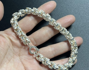 May include: A silver bracelet with a textured, interconnected design. The bracelet is made of small, irregular silver pieces linked together to form a chain. It features a silver clasp. The bracelet is displayed on a hand against a dark background.