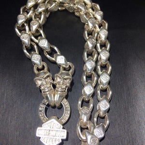 May include: Silver-toned motorcycle chain necklace featuring a skull pendant and Harley-Davidson logo. The chain links have a unique design, and the necklace is displayed against a dark background. The necklace is a bold accessory.