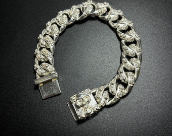 May include: Silver-toned curb chain bracelet with clear, sparkling accents. The bracelet features a skull design on the clasp. The links are thick and embellished with small, round stones. The bracelet is on a dark background.