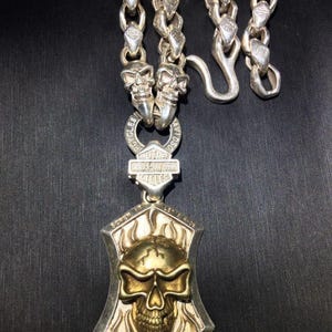 May include: Silver-toned necklace featuring a skull pendant with a gold-colored skull design. The pendant is framed by a shield shape with flame details. The necklace includes a chain with skull accents and a Harley-Davidson logo.