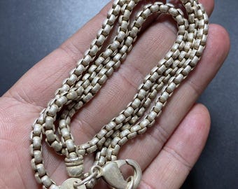 May include: A silver-colored necklace with a box chain design. The necklace has a lobster clasp closure and a small circular tag with the number "925" stamped on it. The chain is made of small, square-shaped links.