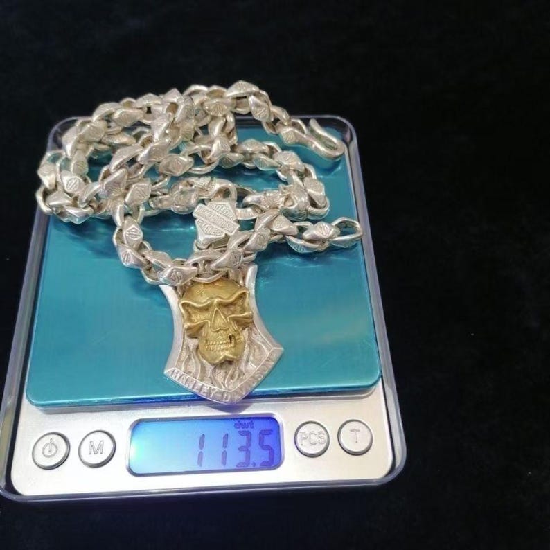 May include: Silver necklace with a skull pendant and a chain link design. The pendant has a gold-colored skull and the words "HARLEY-D" engraved on it. The necklace is on a digital scale, which reads 113.5.