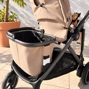 Könnte beinhalten: Ein beigefarbener Kinderwagen mit schwarzem Rahmen, einem großen beigen Aufbewahrungskorb an der Vorderseite und einer passenden beigen Sitzeinheit. Der Kinderwagen verfügt über schwarze Räder, einen schwarzen Schiebegriff und ein kleines Plüschlöwenspielzeug.